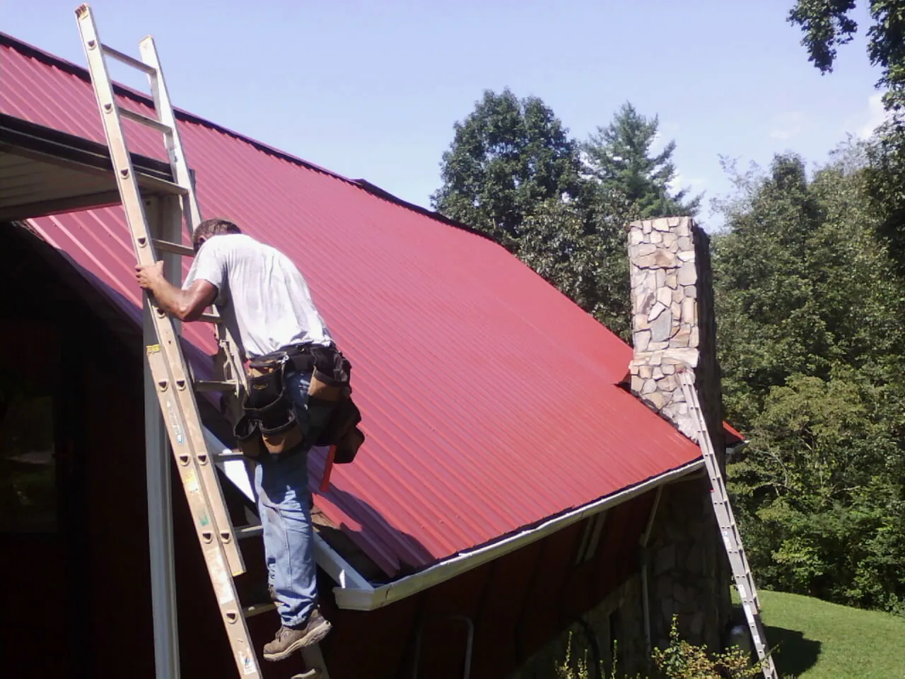 Roofer on a ladder beside a red metal roof and stone chimney — Soffit & Fascia Repair by Roofing Brothers in Lavon